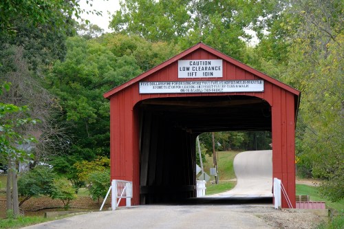 A red covered bridge with a low clearance sign, surrounded by green trees and a winding road.