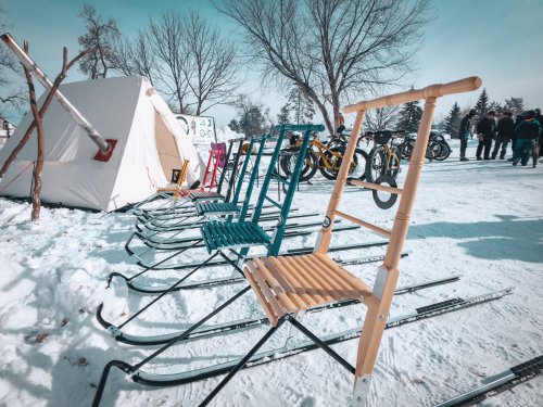 A row of colorful wooden sleds on skis in a snowy landscape, with a tent and people in the background.