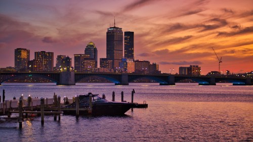 City skyline at sunset with a bridge and a boat docked in the foreground, reflecting vibrant colors in the water.