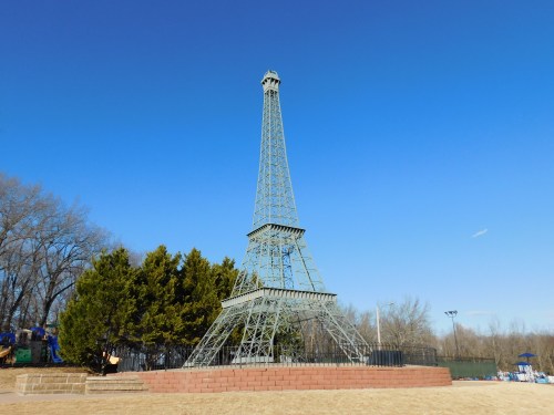 A miniature Eiffel Tower stands against a clear blue sky, surrounded by trees and a sandy area.
