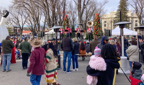 A festive crowd gathers at an outdoor event with musicians performing on stage, surrounded by holiday decorations.