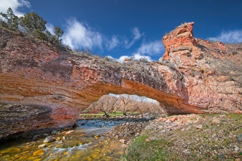 A natural stone arch spans over a clear stream, surrounded by rocky terrain and sparse vegetation under a blue sky.