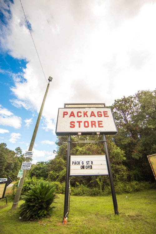 Sign for a package store with "PACKAGE STORE" displayed prominently, surrounded by greenery and a blue sky.