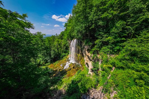 A stunning waterfall cascades down rocky cliffs, surrounded by lush green trees and a clear blue sky.