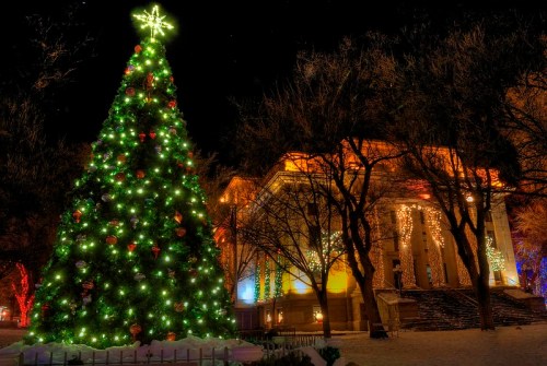 A brightly lit Christmas tree stands in front of a beautifully decorated building at night, surrounded by snow.