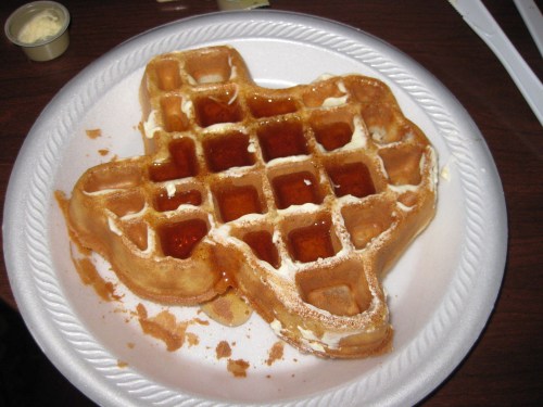 A Texas-shaped waffle drizzled with syrup and topped with a dollop of butter on a paper plate.