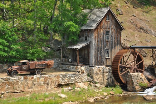 A rustic wooden mill with a water wheel beside an old truck loaded with logs, surrounded by greenery and rocky terrain.
