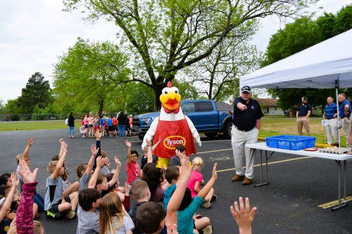 A chicken mascot stands in front of children raising their hands, with adults and a table in the background under a tent.