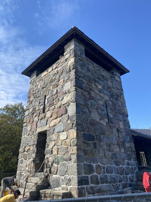 Stone tower with a flat roof against a blue sky, surrounded by trees and people at the base.