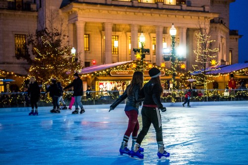 Two skaters glide on an ice rink, surrounded by festive lights and holiday decorations in a lively winter scene.