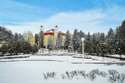 A snow-covered landscape featuring a yellow building with red roofs, surrounded by evergreen trees under a blue sky.