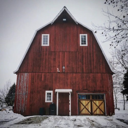 A red barn with a peaked roof, snow-covered ground, and a wooden door, set against a gray winter sky.