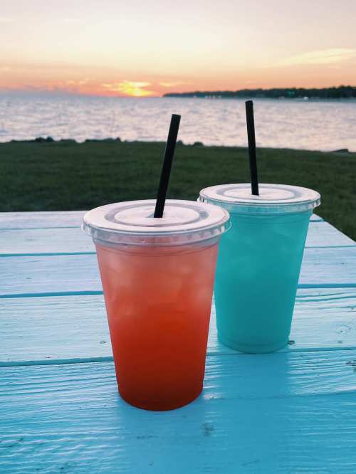 Two colorful drinks in clear cups sit on a blue table, with a sunset over the water in the background.