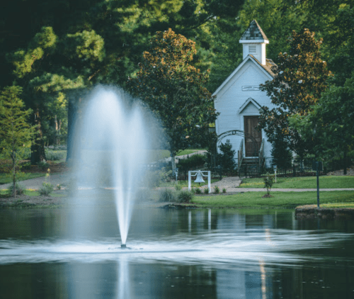 A serene park scene featuring a fountain in a pond, with a quaint white building and lush greenery in the background.