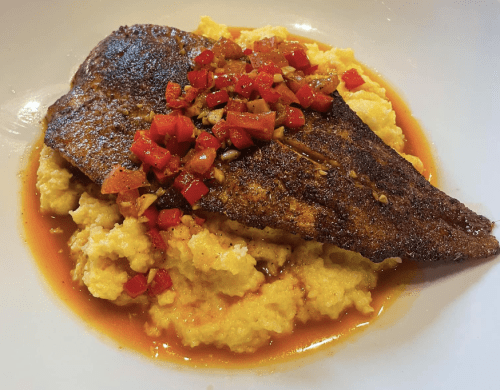 A plate of blackened fish atop creamy grits, garnished with diced tomatoes and peppers in a savory sauce.