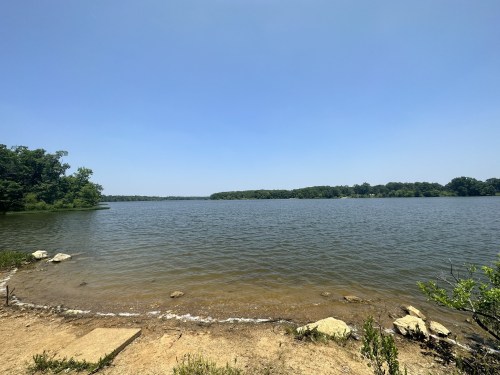 A serene lake view with clear blue skies, surrounded by greenery and rocky shoreline.