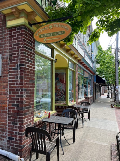 A cozy café with outdoor seating, colorful signage, and large windows, set on a tree-lined street.