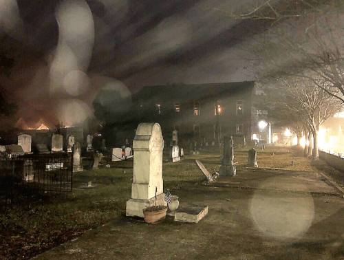 A foggy cemetery at night, featuring gravestones and a dimly lit building in the background.