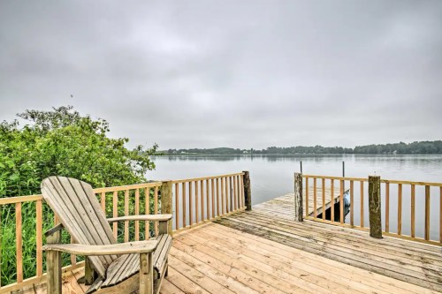 A wooden deck with an Adirondack chair overlooks a calm lake under a cloudy sky.
