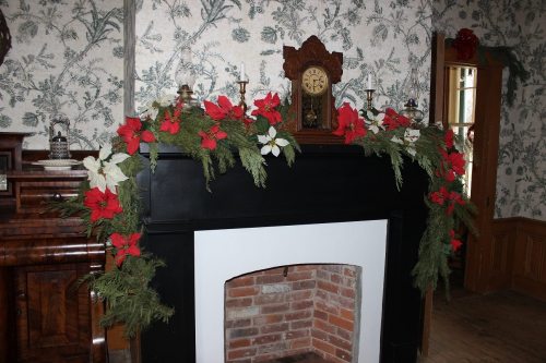 A decorated fireplace with a garland of red and white flowers, set against floral wallpaper and a vintage clock.