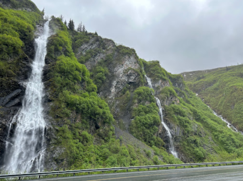 Two waterfalls cascade down a lush, green mountainside under a cloudy sky, alongside a winding road.