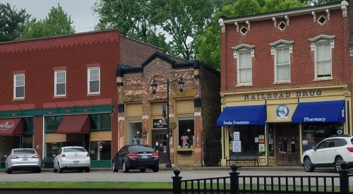 Historic buildings line a street, featuring shops like a pharmacy and a soda fountain, with parked cars in front.