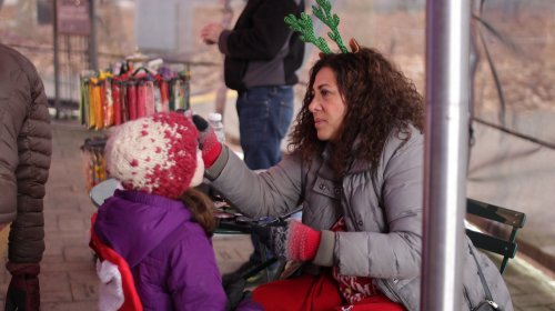 A woman with reindeer antlers applies face paint to a child wearing a winter hat at an outdoor event.