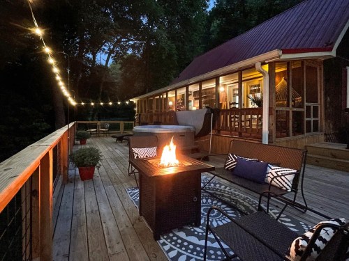 Cozy outdoor deck at night with a fire pit, string lights, and a view of a cabin surrounded by trees.