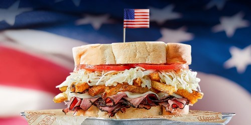 A towering sandwich with layers of roast beef, fries, lettuce, and tomato, topped with a small American flag, against a flag backdrop.