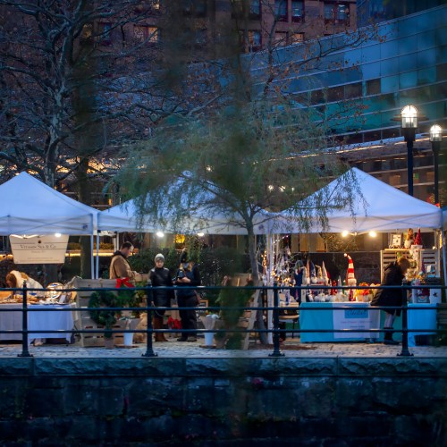 A festive market with white tents, vendors, and holiday decorations along a waterfront, illuminated by soft lights.