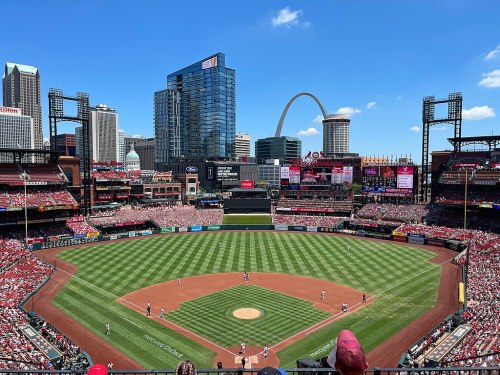 A panoramic view of a baseball stadium filled with fans, featuring a city skyline and an iconic arch in the background.