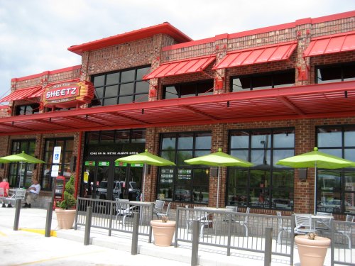 Exterior of a Sheetz convenience store with a red roof, large windows, and outdoor seating under green umbrellas.