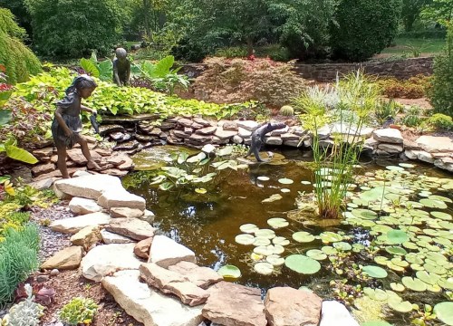 A serene garden scene featuring a pond with lily pads and two sculptures of children playing near the water's edge.