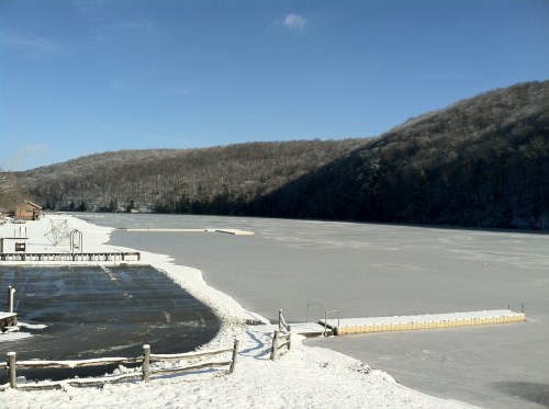 A frozen lake surrounded by snow-covered hills under a clear blue sky, with a dock and parking area visible.