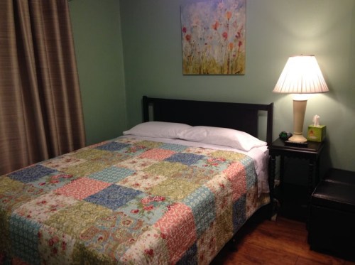 Cozy bedroom with a patchwork quilt, bedside lamp, and floral artwork on the wall. Warm, inviting atmosphere.
