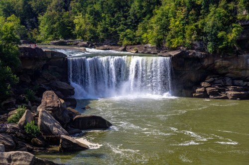 A scenic waterfall cascading over rocks into a green river, surrounded by lush trees and a clear blue sky.