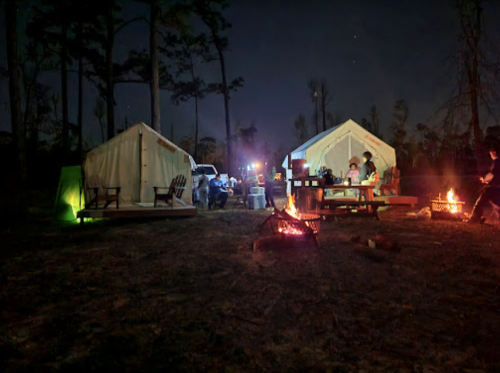 Two glamping tents at night, surrounded by campfires and trees, with people enjoying the outdoor setting.