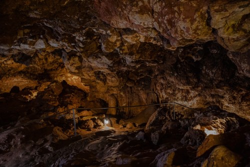 Dimly lit cave interior with rocky formations and a railing, showcasing natural textures and shadows.
