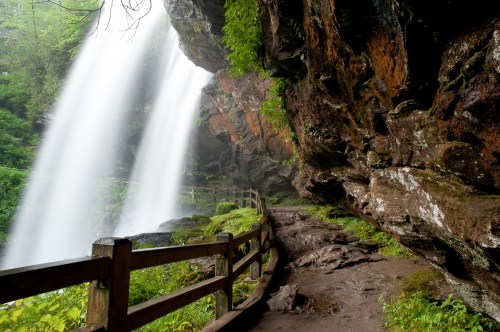 A serene waterfall cascades down rocky cliffs, surrounded by lush greenery and a winding path with a wooden railing.