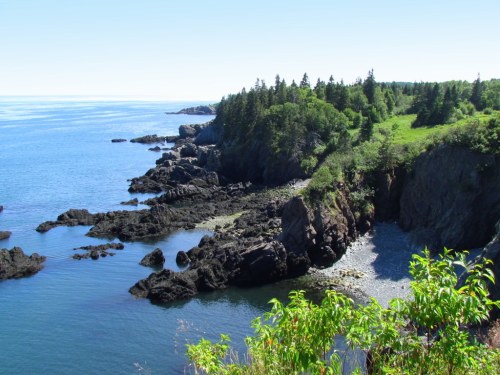A scenic coastal view featuring rocky cliffs, lush greenery, and calm blue waters under a clear sky.