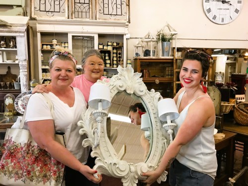 Three women hold a large, ornate mirror in a vintage shop, surrounded by various home decor items.