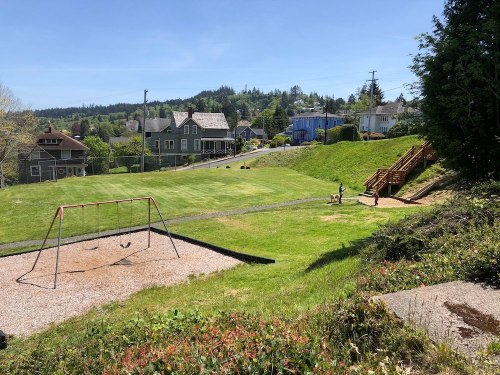 A sunny park with a swing set, grassy area, and houses in the background, featuring a child playing on the grass.