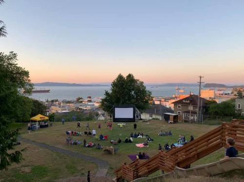Outdoor movie screening on a grassy hill, with people seated and a view of the water and sunset in the background.