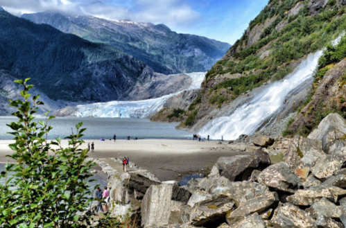 A scenic view of a glacier with a waterfall, surrounded by mountains and people on a sandy beach.