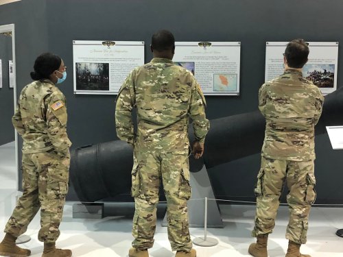 Three soldiers in military uniforms observe historical displays about warfare, with a large cannon in the foreground.