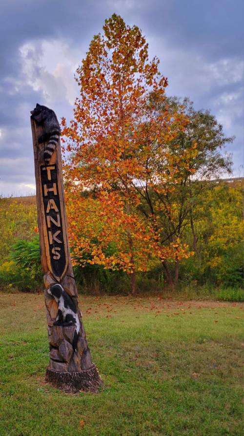A carved wooden post with "Thanks" and animal designs, set against a backdrop of autumn trees and cloudy skies.
