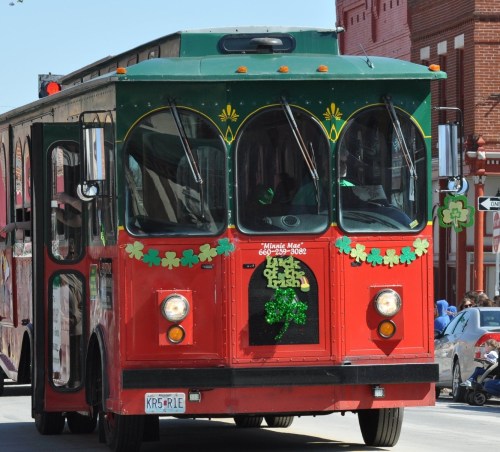 A festive red trolley decorated with green shamrocks drives down a city street on a sunny day.