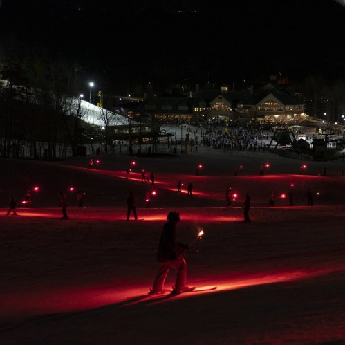 Skiers with red lights glide down a snowy slope at night, with a bustling lodge illuminated in the background.