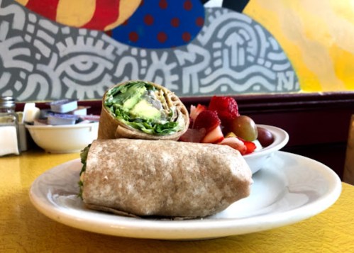 A whole grain wrap filled with greens, served with a side of mixed fruit on a yellow table.