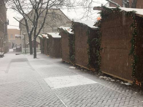 Snow falls on a row of wooden market stalls decorated with lights and greenery, set in a quiet, wintry street.